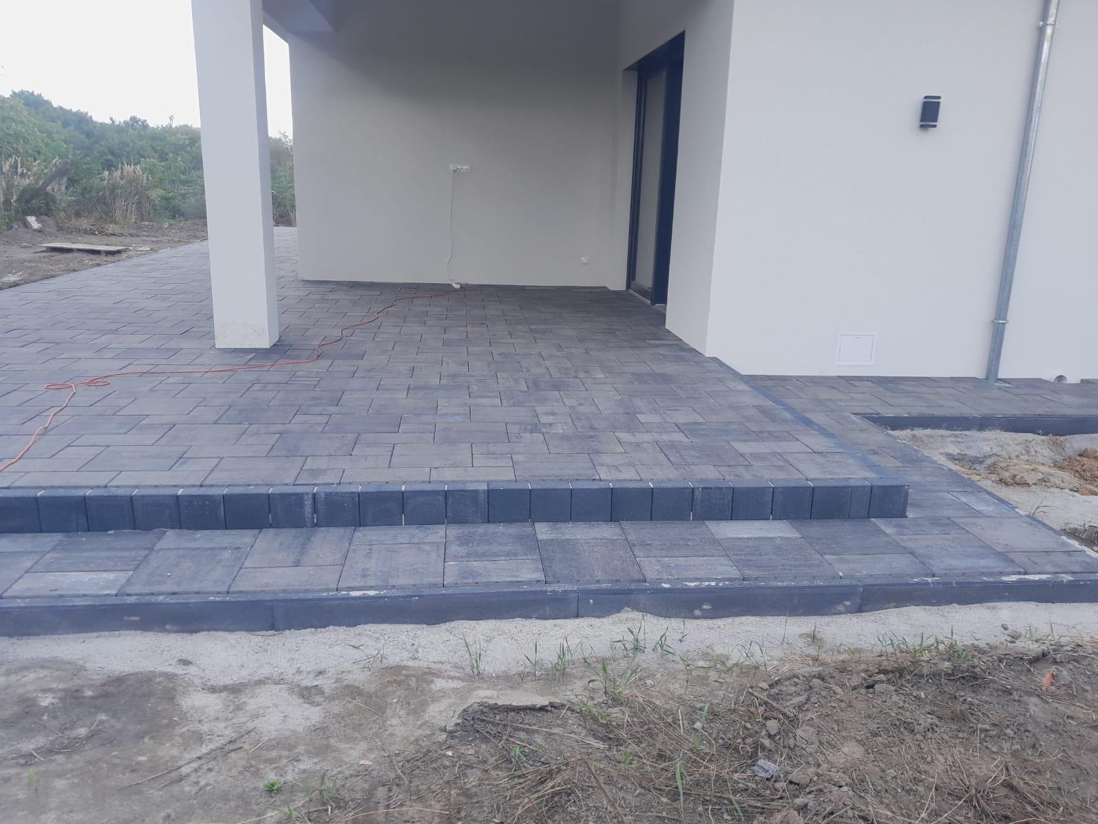 Ułożona kostka brukowa w odcieniach szarości tworząca taras z dwoma stopniami przed nowoczesnym domem, widoczne niedokończone prace ziemne obok.