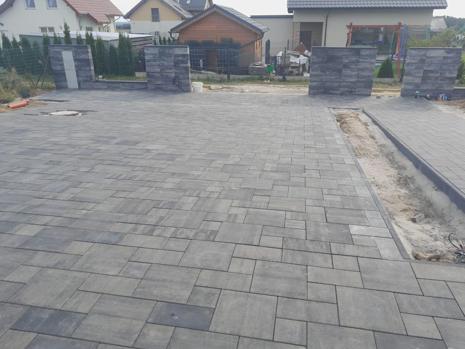 Ułożona szara kostka brukowa na podjeździe z elementami imitującymi kamień, widoczne słupki z tego samego materiału, w tle zabudowa jednorodzinna.