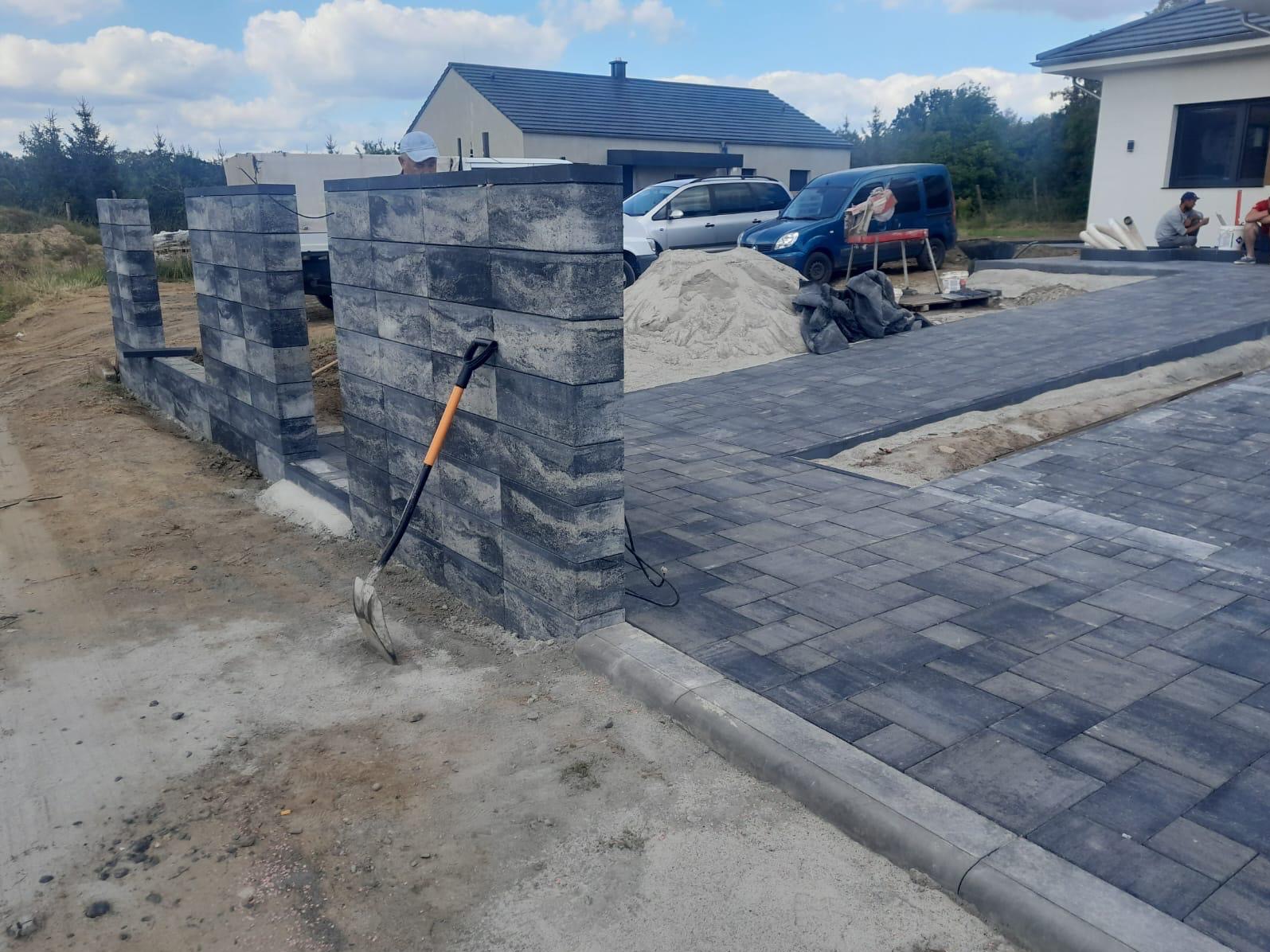 Fragment posesji w trakcie prac: wznoszony mur z szarej, ciosanej kostki betonowej, ułożona szara kostka brukowa, widoczna łopata oparta o mur, w tle domy i samochody.
