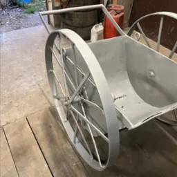 Metalowa taczka dwukołowa, świeżo pomalowana szarą farbą, stoi na drewnianej podłodze w warsztacie, w tle widoczne opony i pojemniki.