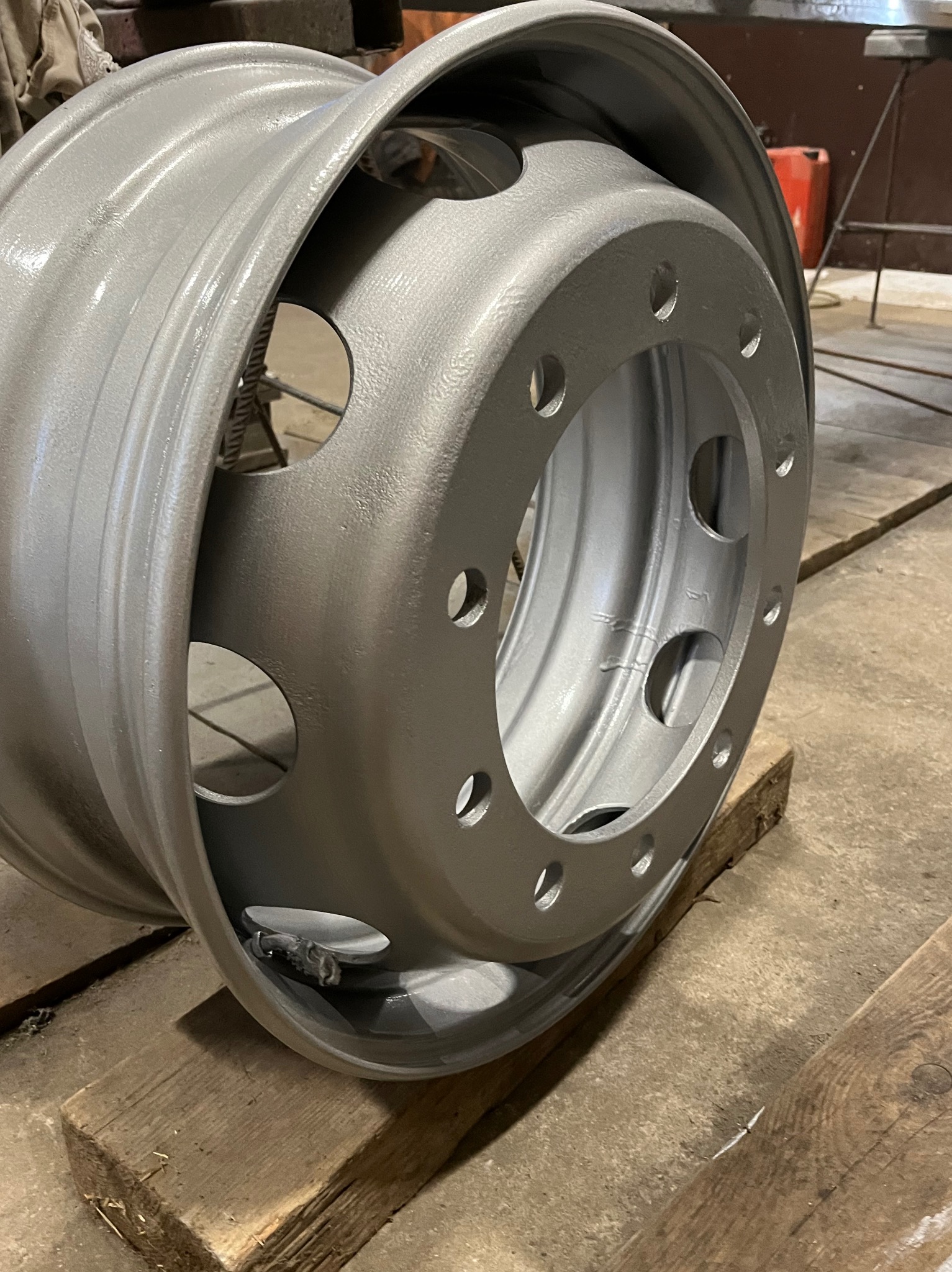 Pomalowana na srebrno felga ciężarówki, oparta na drewnianych klockach w warsztacie, widoczne otwory montażowe i wentylacyjne.