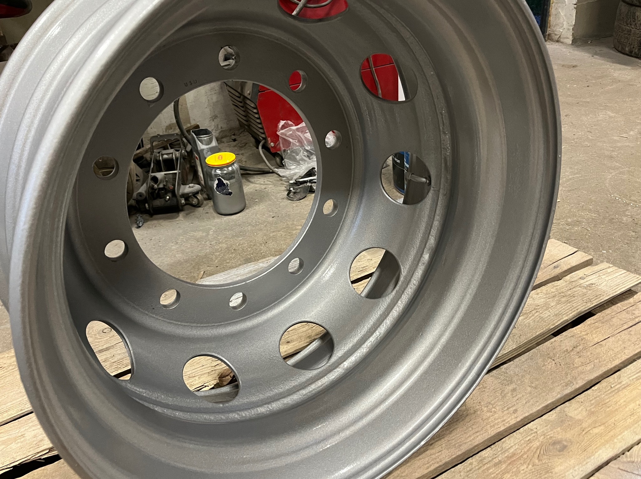 Pomalowana na szaro felga ciężarowa leży na drewnianej palecie. W tle widoczny podnośnik, spawarka i słoik z żółtą nakrętką.