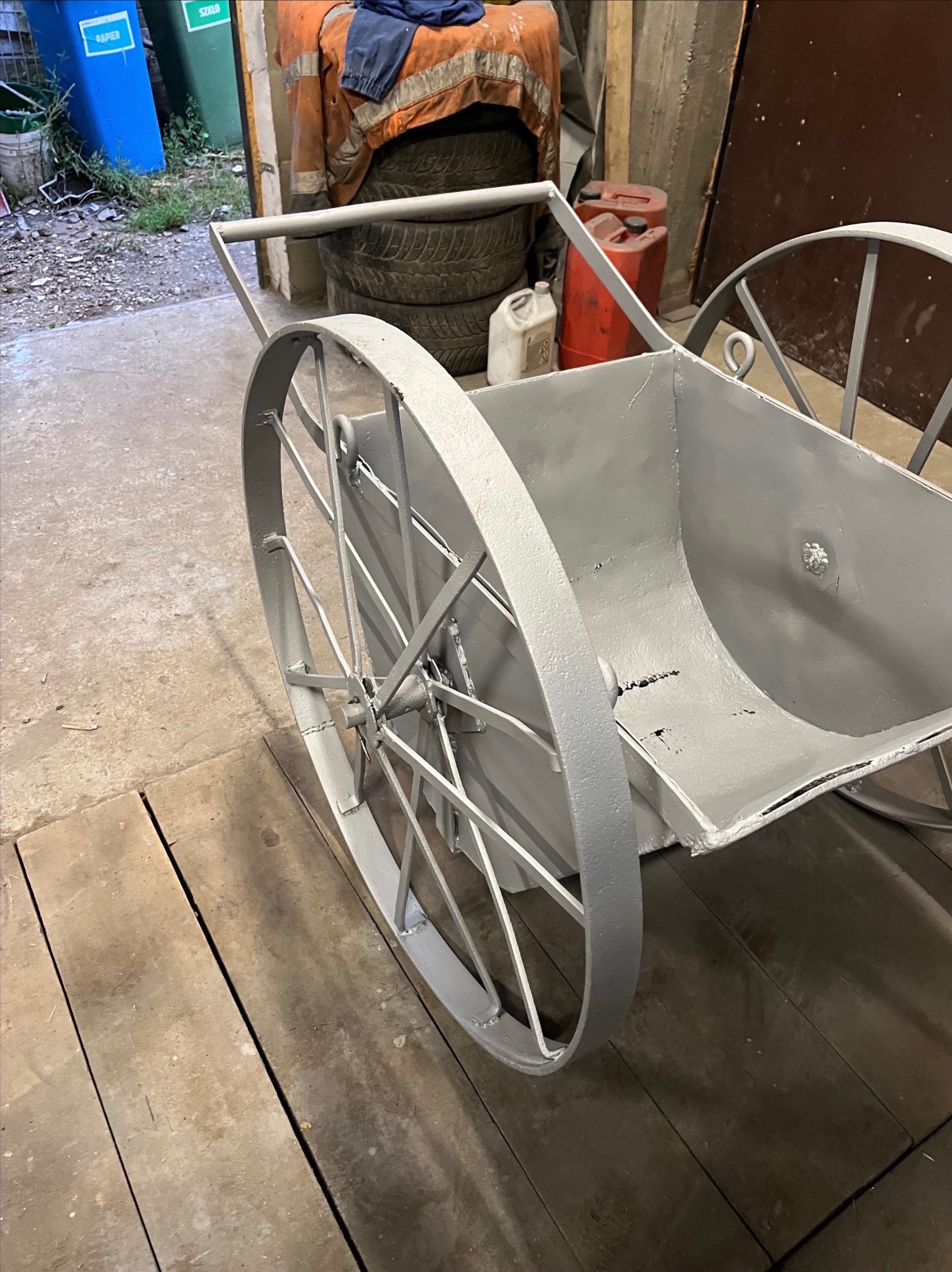 Metalowa taczka dwukołowa, świeżo pomalowana szarą farbą, stoi na drewnianej podłodze w warsztacie, w tle widoczne opony i pojemniki.