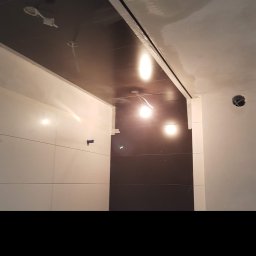 Firma remontowo- budowlana Łukasz Dziurleja - Wnętrze łazienki w trakcie prac wykończeniowych, widoczne białe płytki na ścianie, ciemny sufit podwieszany z instalacją oświetleniową oraz surowy tynk na górze ścian.