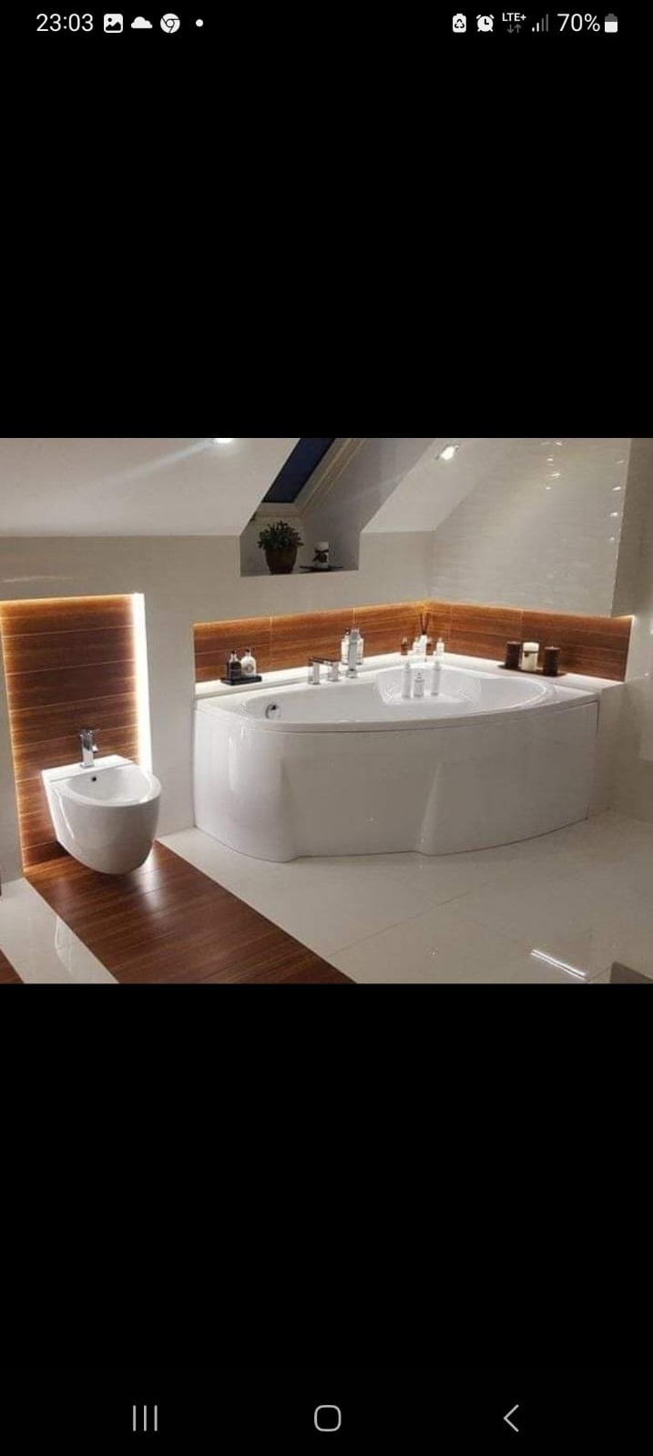 Nowoczesna łazienka na poddaszu z wolnostojącą wanną narożną, bidetem i drewnianymi akcentami podświetlonymi LED, uzupełniona oświetleniem naturalnym z okna dachowego.