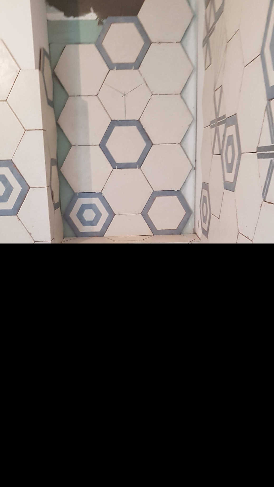 Fragment ściany wyłożonej białymi, heksagonalnymi płytkami z szarym obramowaniem, widoczne nierówne fugi i fragment surowej ściany w górnym rogu.