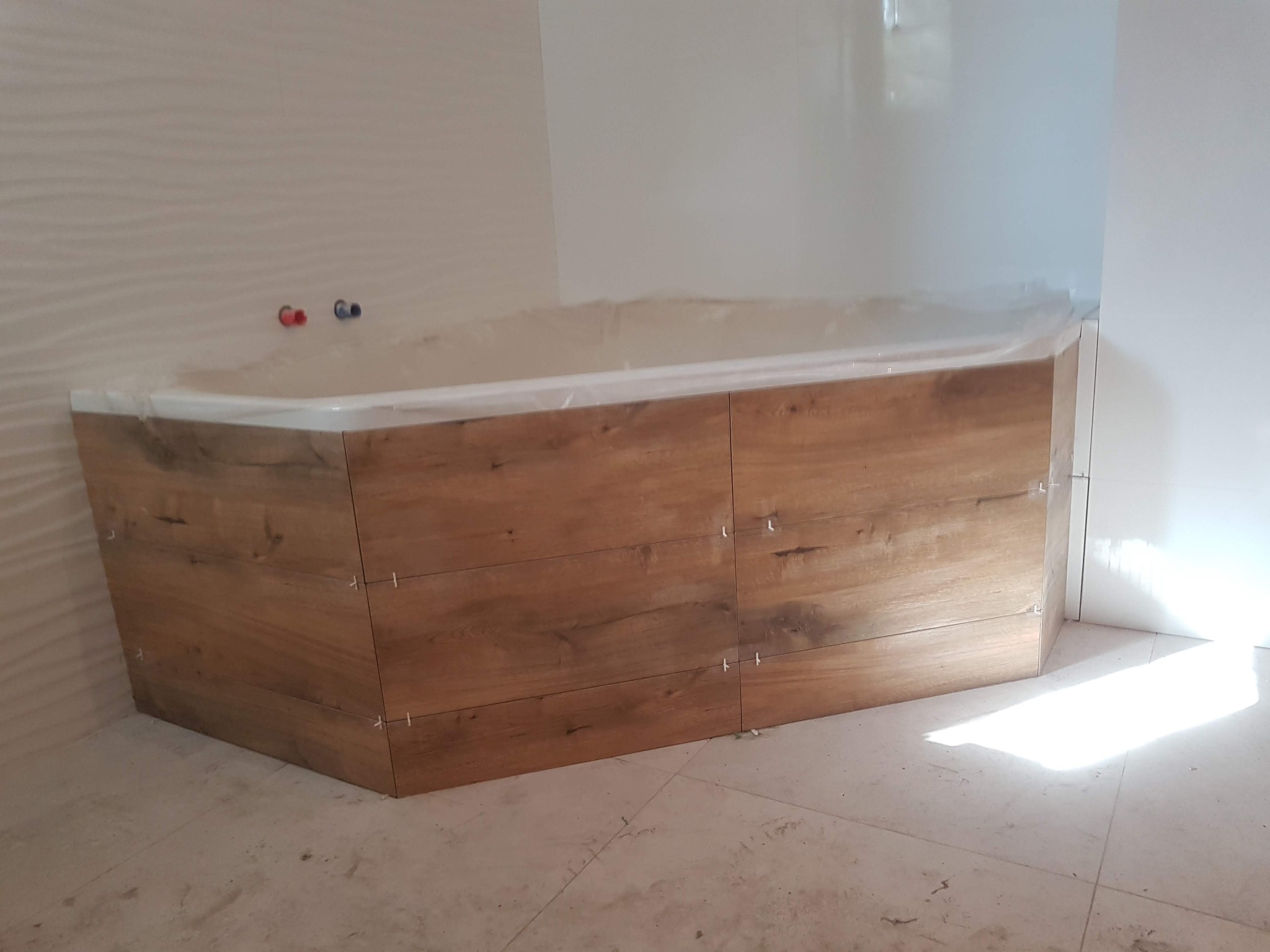 Obudowa wanny wykończona płytkami imitującymi drewno, widoczne krzyżyki dystansowe, ściany częściowo wykończone, podłoga w trakcie prac.