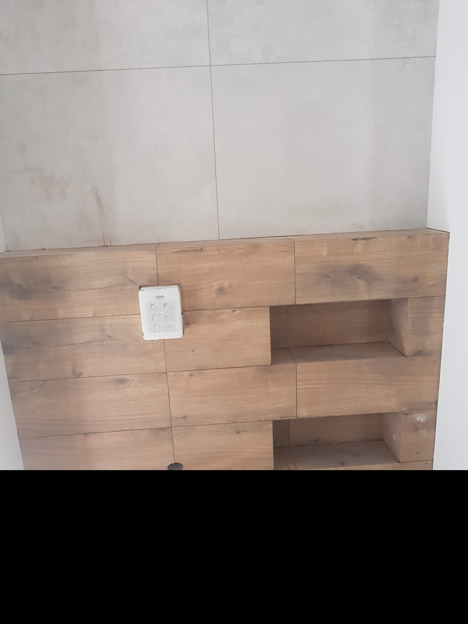 Ściana łazienki wykończona płytkami imitującymi drewno z wnękami i panelem sterowania Grohe, górna część ściany wyłożona szarymi płytkami.