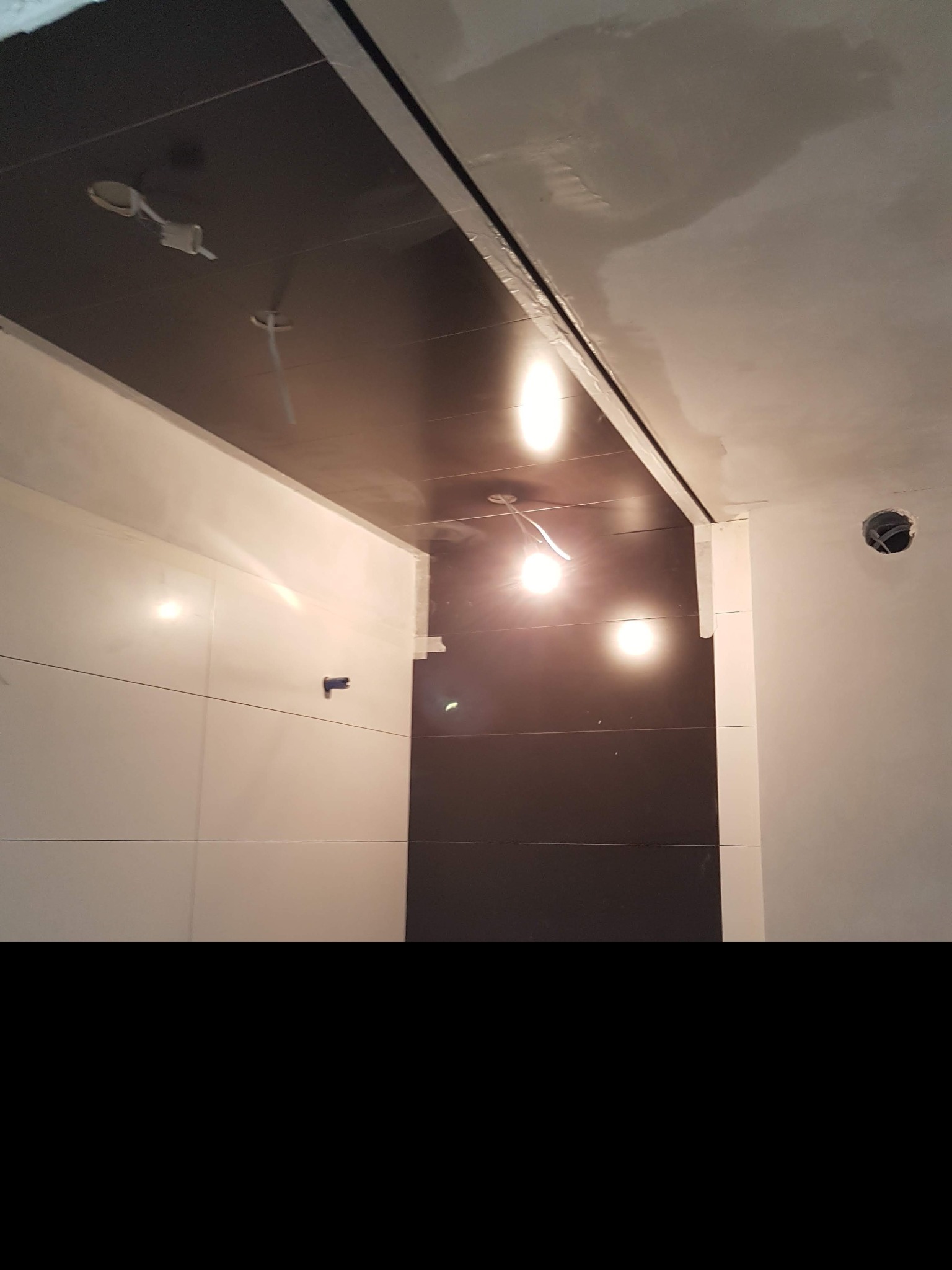 Wnętrze łazienki w trakcie prac wykończeniowych, widoczne białe płytki na ścianie, ciemny sufit podwieszany z instalacją oświetleniową oraz surowy tynk na górze ścian.