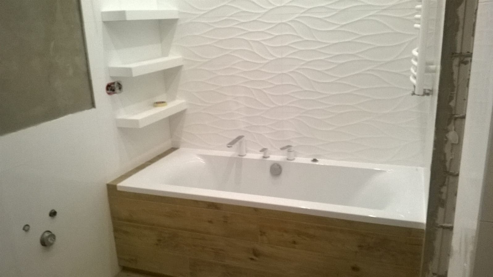 Biała wanna obudowana panelami imitującymi drewno, z baterią i ozdobną, falistą glazurą na ścianie, uzupełniona o białe półki wnękowe. Widoczne ślady prac remontowych na ścianie po prawej.