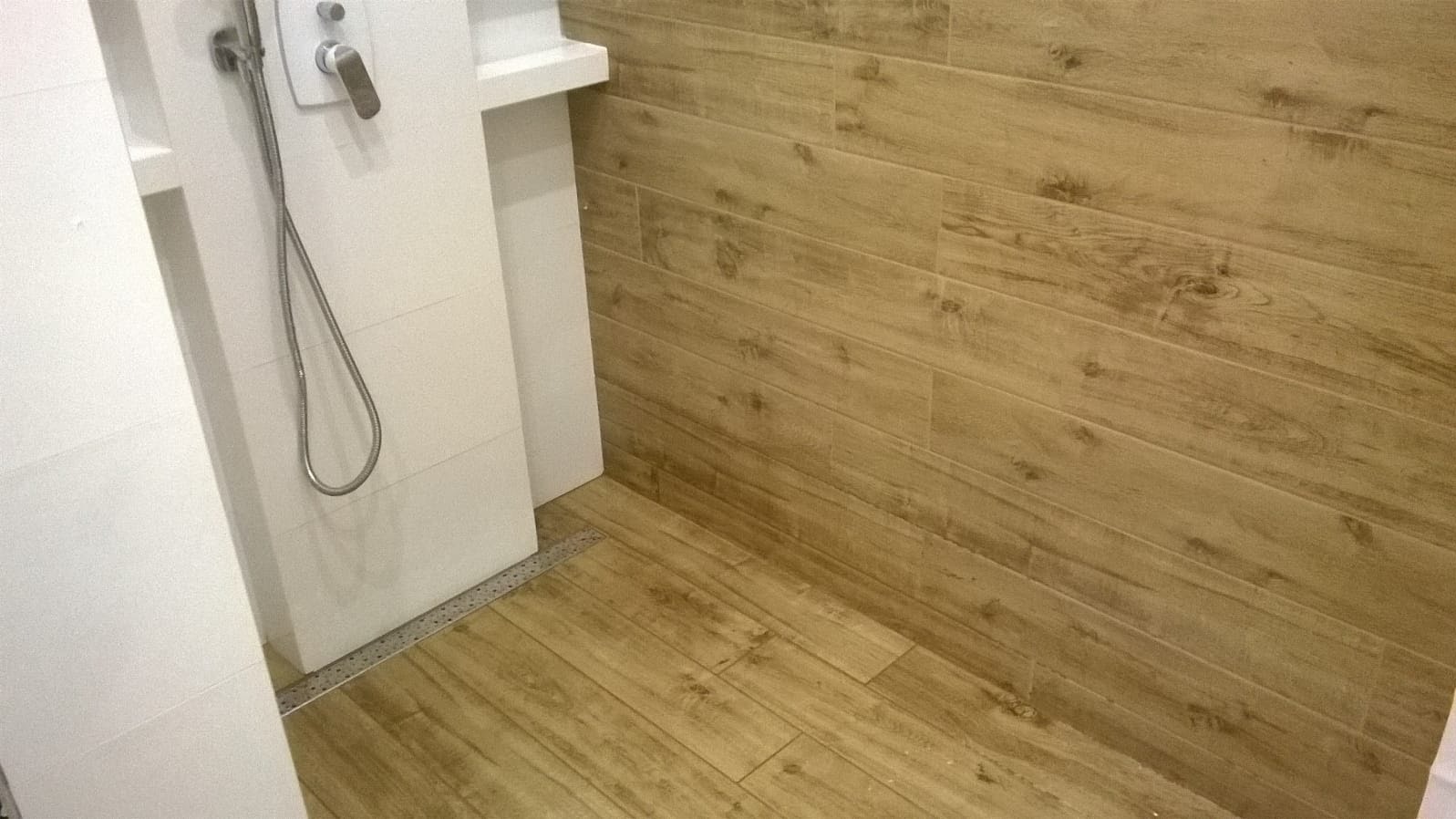 Ukończona łazienka z drewnopodobnymi płytkami na podłodze i ścianie, kabina prysznicowa z odpływem liniowym, chromowana bateria i słuchawka prysznicowa.