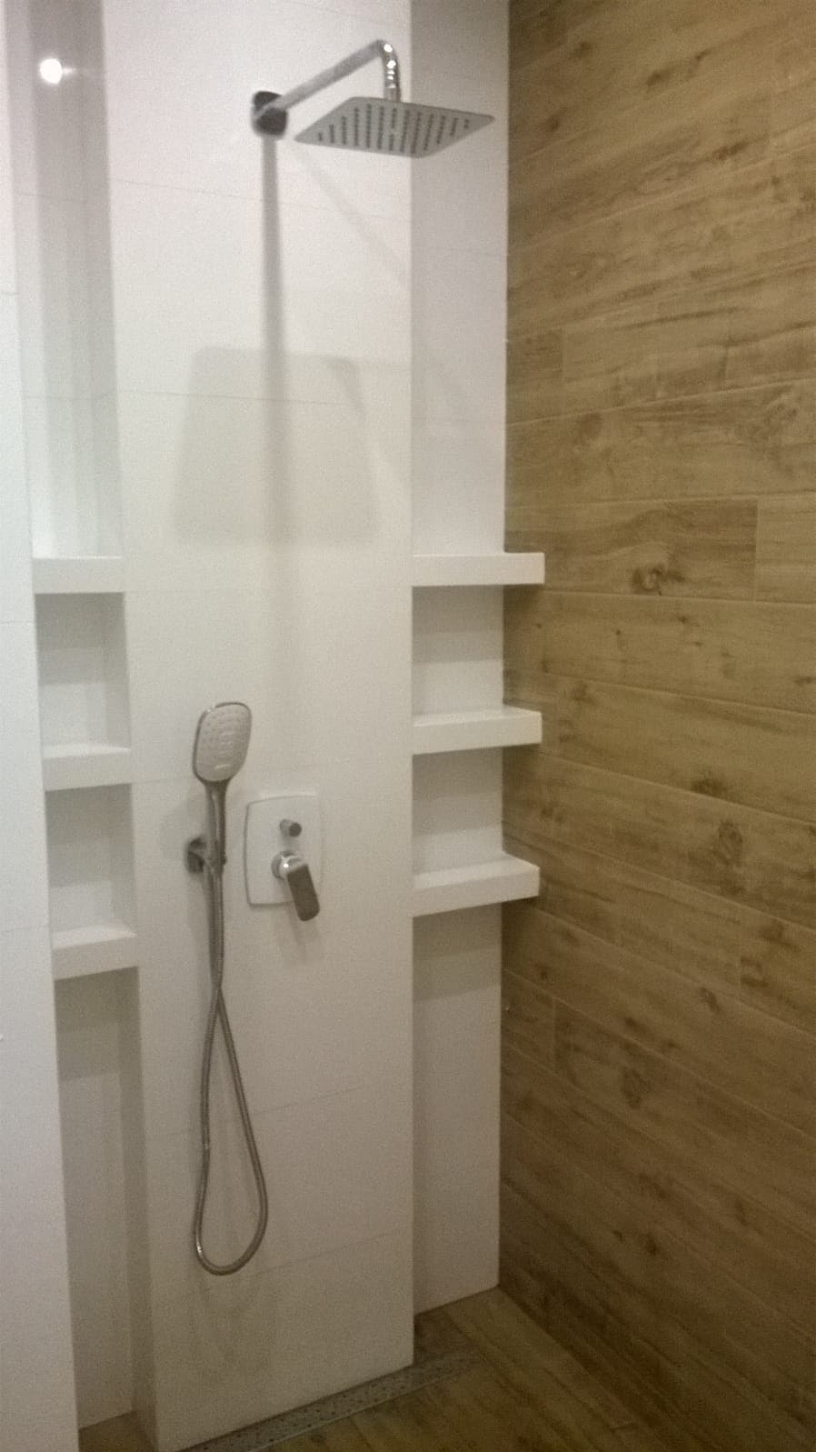 Kabina prysznicowa z deszczownicą i słuchawką, wykończona białymi i drewnopodobnymi płytkami, z wnękami pełniącymi rolę półek.