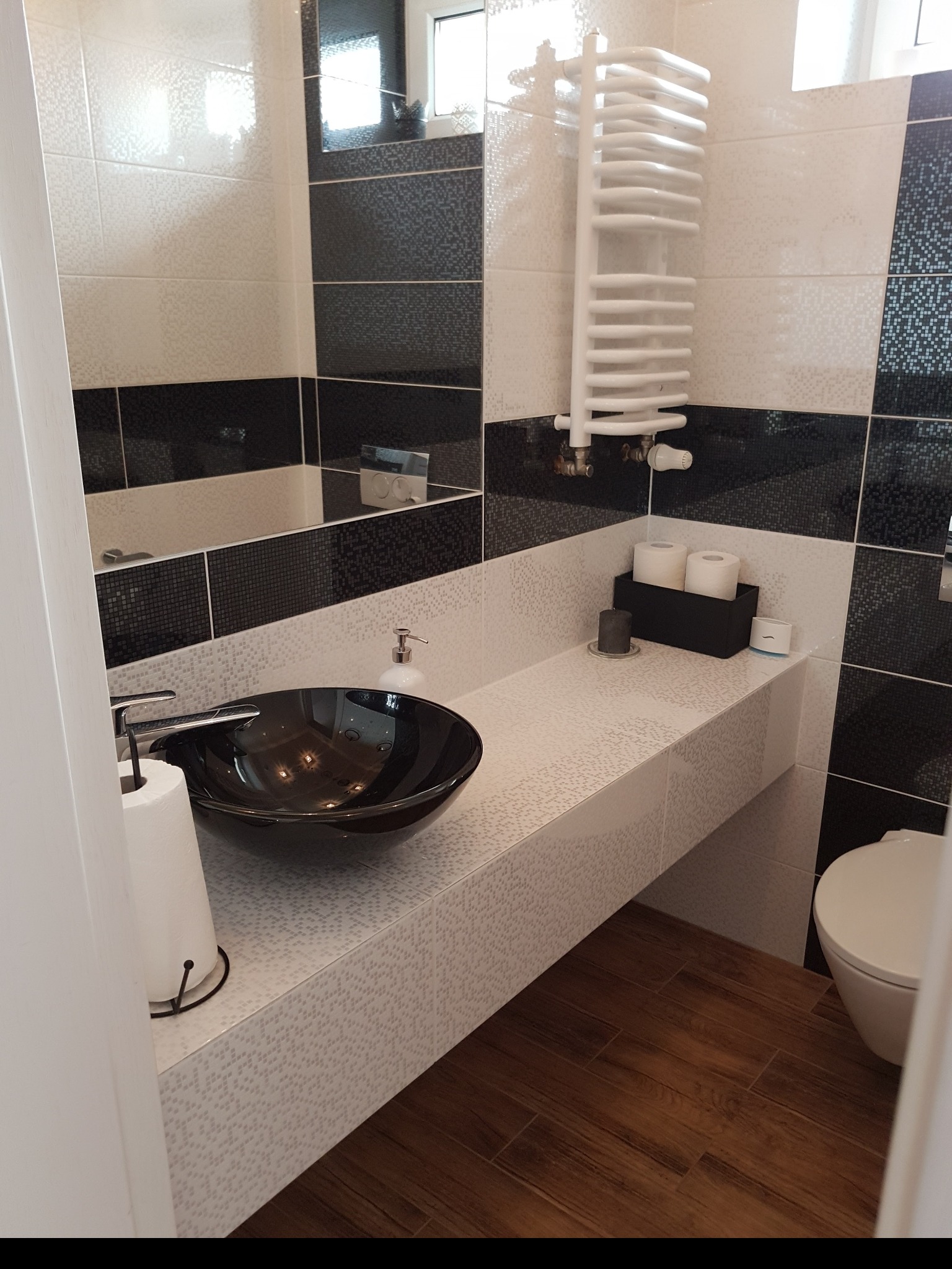 Nowoczesna łazienka z czarno-białymi mozaikowymi płytkami, okrągłą, czarną umywalką na blacie, grzejnikiem drabinkowym i drewnopodobnymi płytkami podłogowymi.