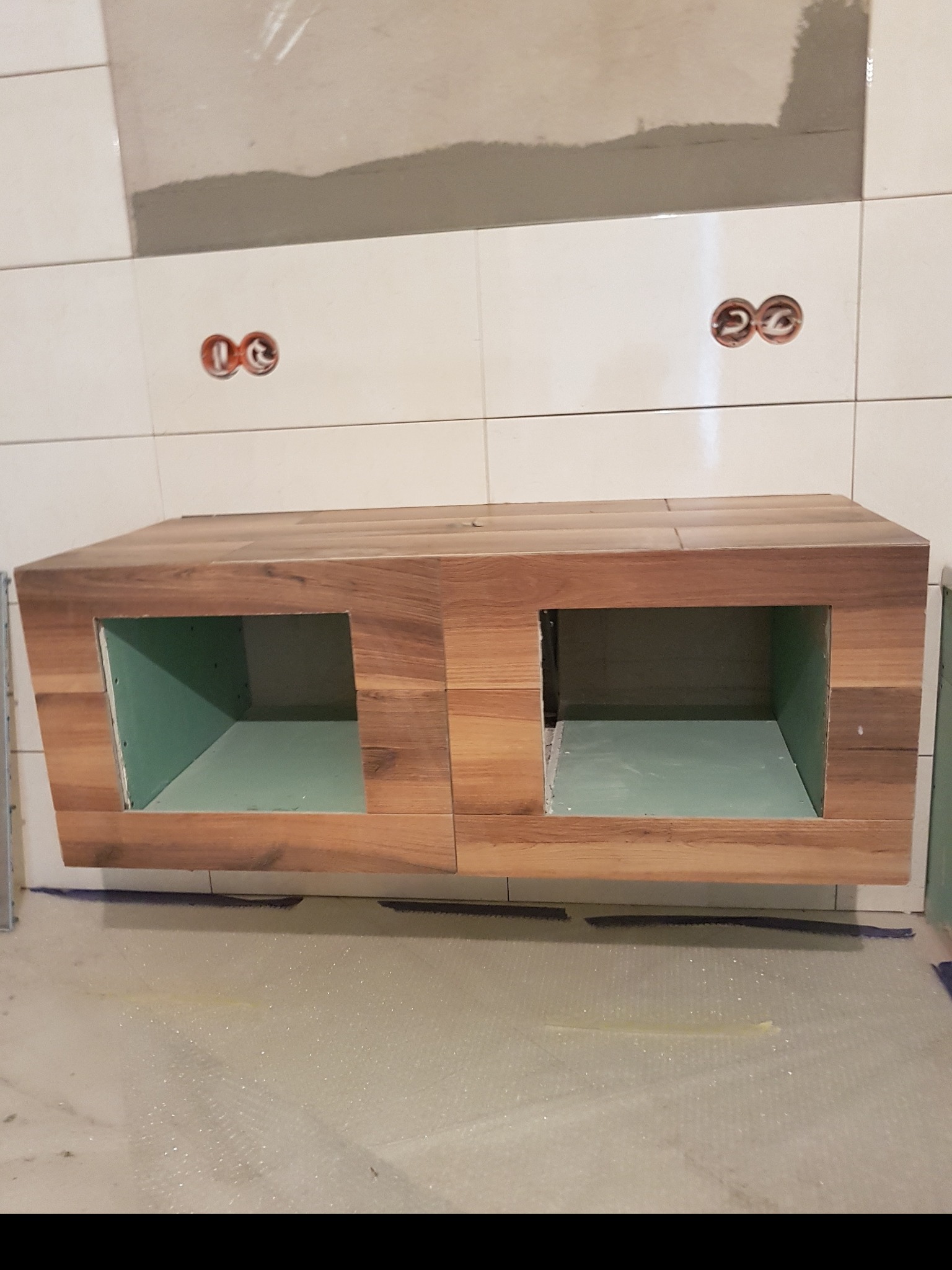 Drewniana szafka pod umywalkę, w trakcie montażu, z widocznymi otworami na rury i zielonym wnętrzem, zamontowana na białych płytkach z widocznymi gniazdkami elektrycznymi.