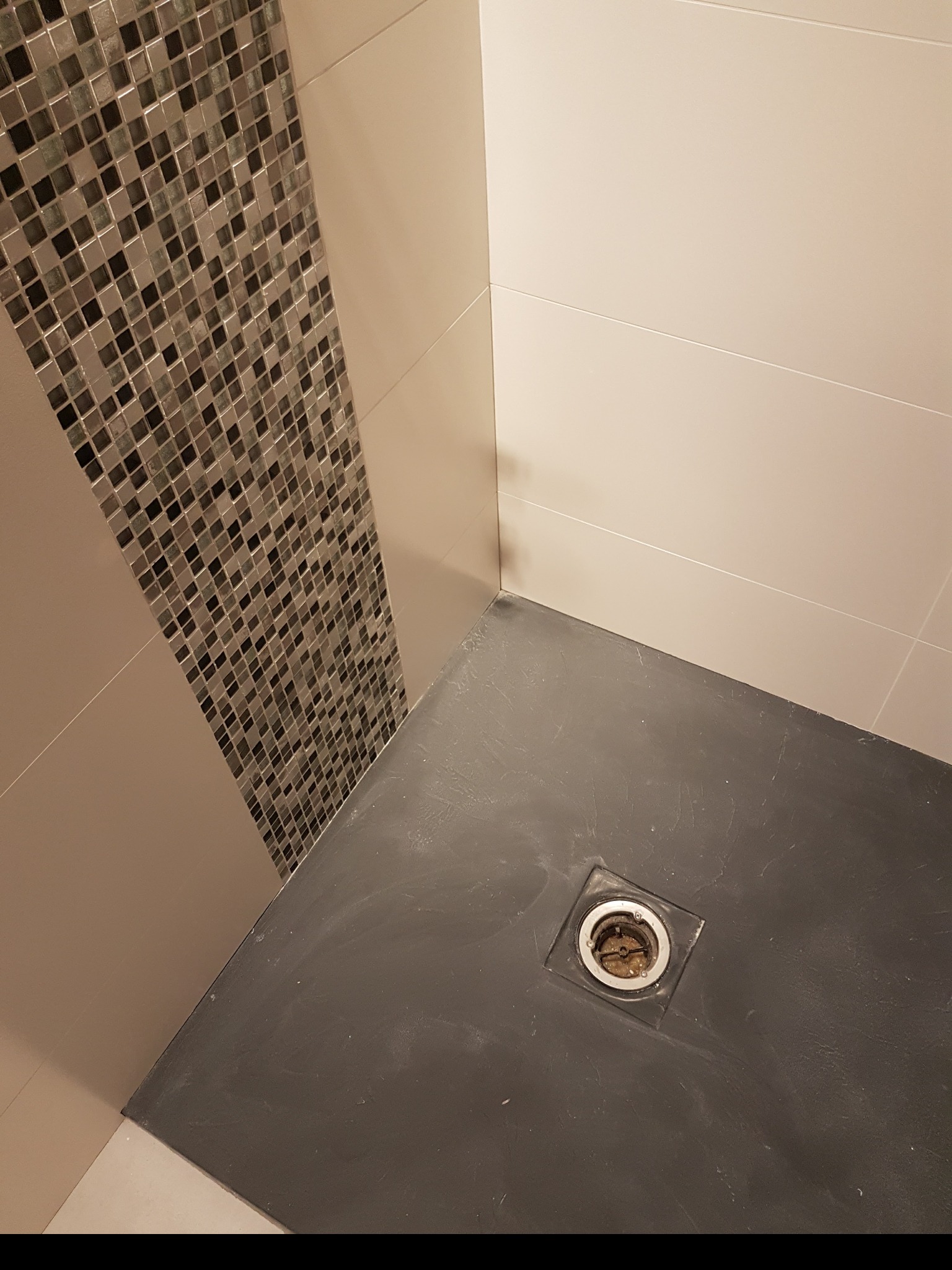 Nowoczesny odpływ liniowy w ciemnym brodziku, obok ściana z beżowych płytek i pionowy pas mozaiki w odcieniach szarości i srebra.