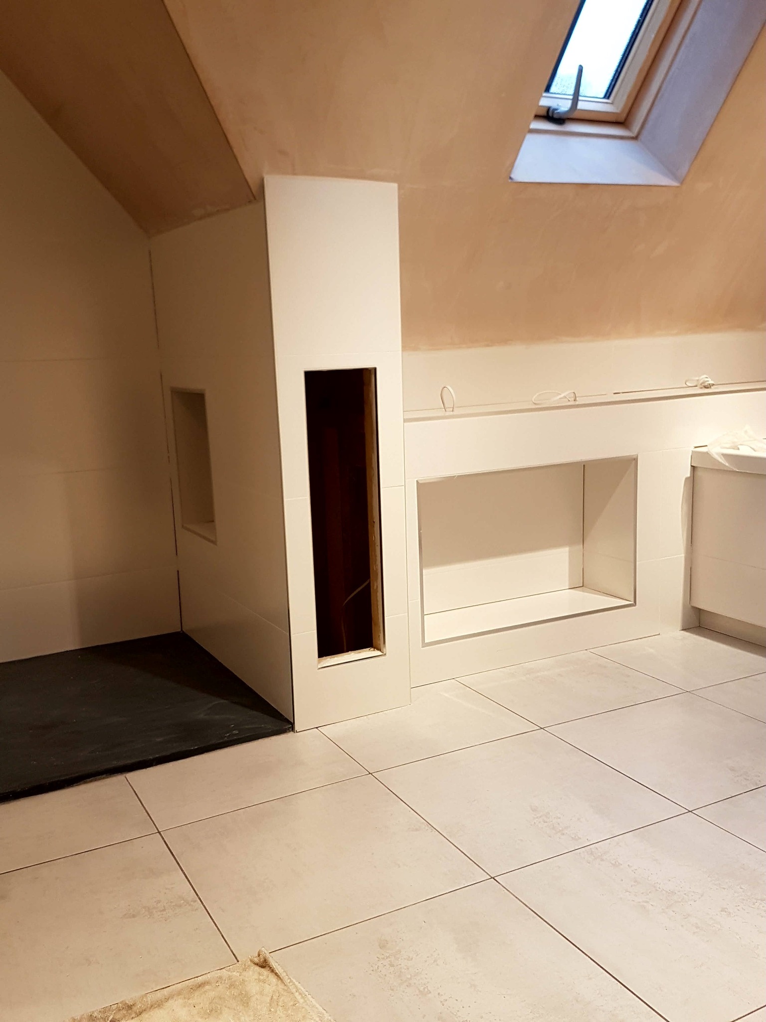 Łazienka w trakcie remontu: białe płytki na podłodze i ścianach, zabudowa pod umywalkę z otworami, widoczne okno dachowe i surowy strop.