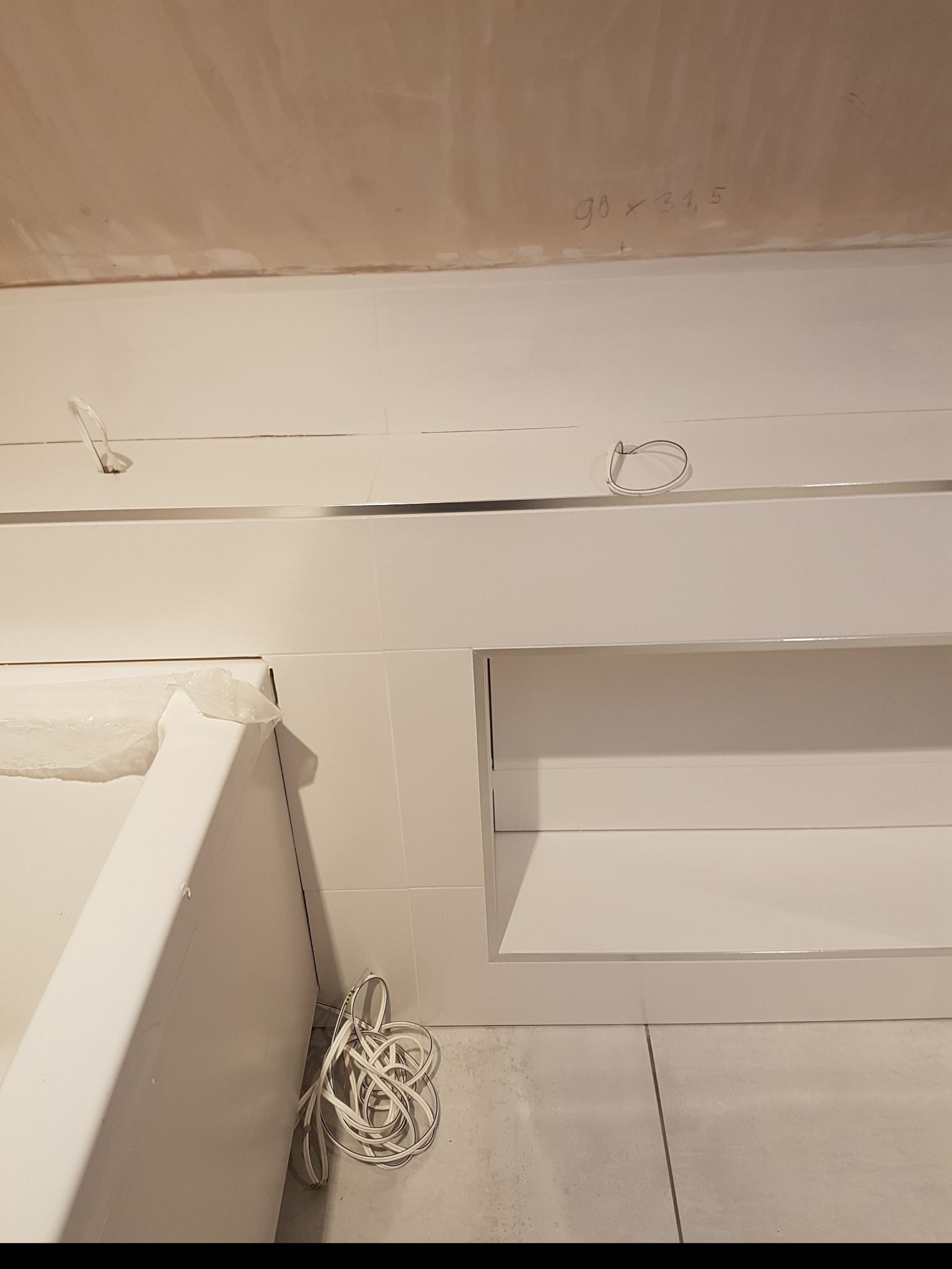 Białe płytki ścienne z metalową listwą, wnęka w ścianie, fragment wanny zabezpieczony folią, zwój kabli na podłodze, surowy sufit z wymiarami zapisanymi ołówkiem.