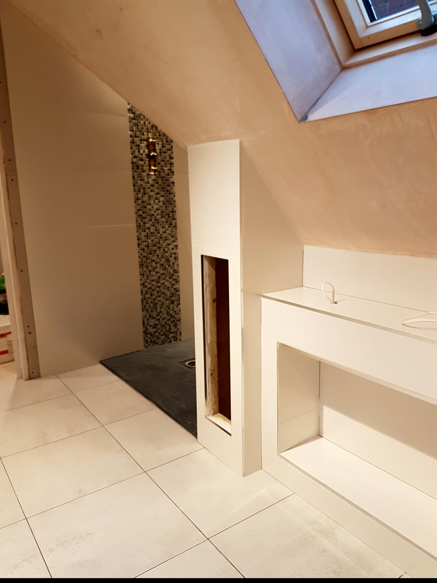 Łazienka na poddaszu w trakcie prac wykończeniowych: białe płytki podłogowe, kabina prysznicowa z mozaiką, zabudowa z półkami, okno dachowe i ściany przygotowane do malowania.