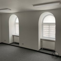PRZEDSIĘBIORSTWO OGÓLNOBUDOWLANE KOZBUD SPÓŁKA Z OGRANICZONĄ ODPOWIEDZIALNOŚCIĄ - Wnętrze biura z dwoma oknami w kształcie łuku, świeżo pomalowane ściany na biało, szary dywan i białe grzejniki.