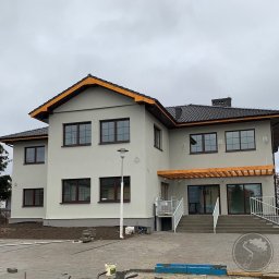 PRZEDSIĘBIORSTWO OGÓLNOBUDOWLANE KOZBUD SPÓŁKA Z OGRANICZONĄ ODPOWIEDZIALNOŚCIĄ - Dwupiętrowy dom po remoncie elewacji w odcieniach szarości z ciemnobrązowymi ramami okiennymi i drewnianymi elementami wykończeniowymi dachu oraz zadaszenia nad wejściem, widoczny z poziomu...