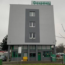 PRZEDSIĘBIORSTWO OGÓLNOBUDOWLANE KOZBUD SPÓŁKA Z OGRANICZONĄ ODPOWIEDZIALNOŚCIĄ - Szary budynek biurowy z zielonymi akcentami i napisem 'Gazomet' na dachu, widziany z poziomu ulicy w pochmurny dzień.