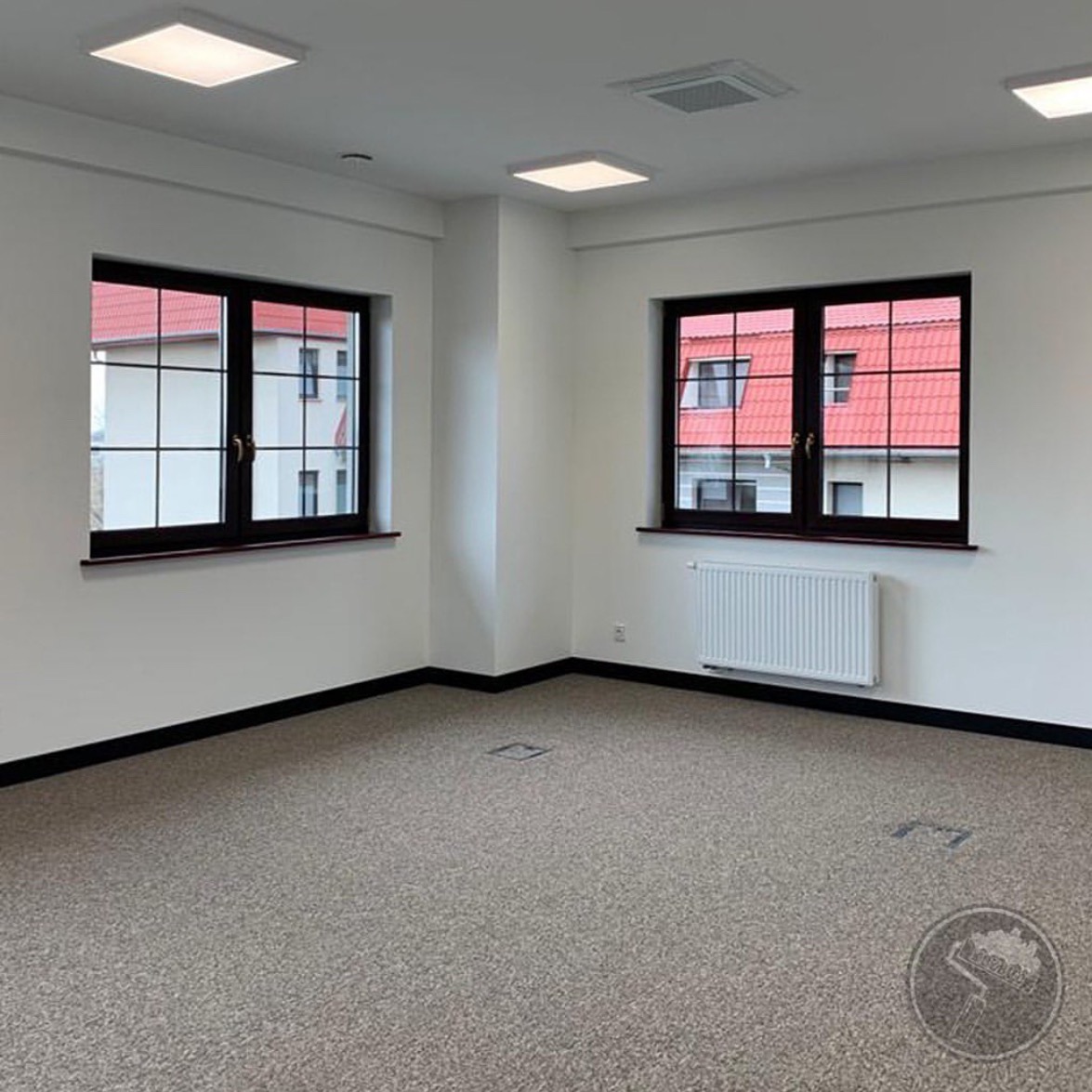 Puste, jasne pomieszczenie biurowe z dwoma oknami w ciemnych ramach, szarym dywanem w plamki i białymi ścianami oraz grzejnikiem pod oknem.