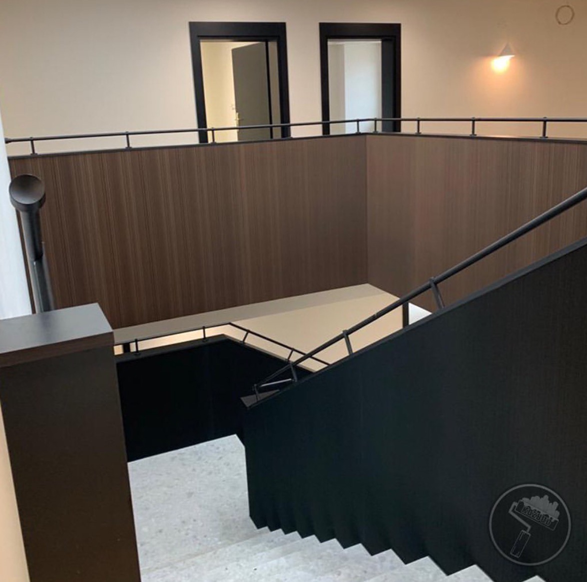 Wnętrze klatki schodowej w budynku biurowym, widok z góry na schody z czarną balustradą i szarymi stopniami, ściany wykończone panelami w odcieniach brązu, dwie pary drzwi z czarnymi ościeżnicami...