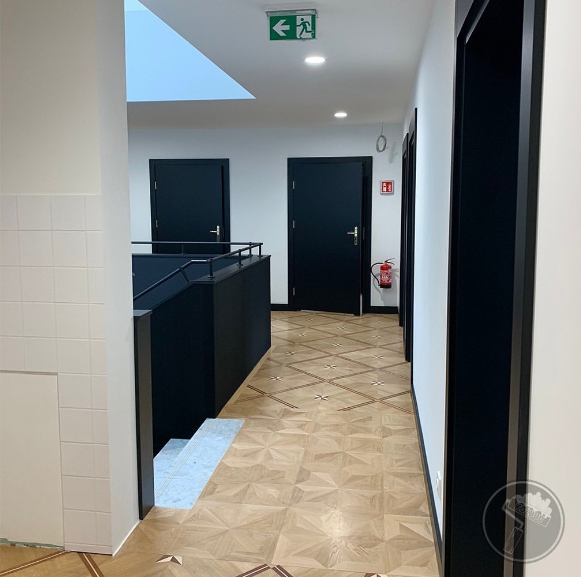 Korytarz biurowy po remoncie: parkiet w geometryczny wzór, czarne drzwi i ramy, białe ściany, widoczny fragment schodów i sufit z oknem dachowym.
