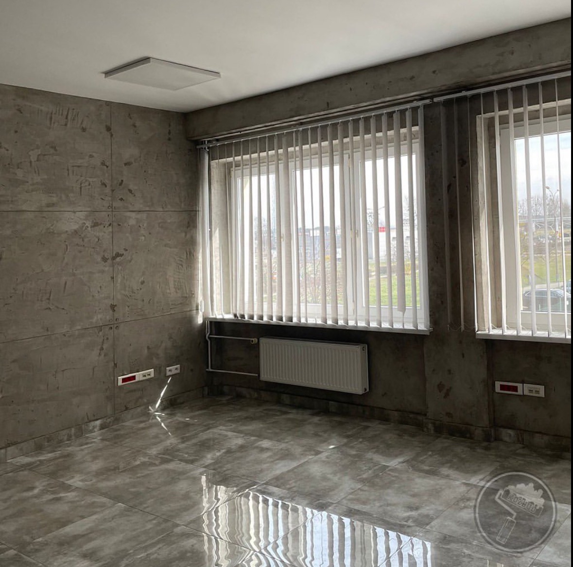 Wnętrze wykończonego pokoju biurowego z szarą, betonową fakturą na ścianach i podłodze, pionowe żaluzje w oknach i biały grzejnik.