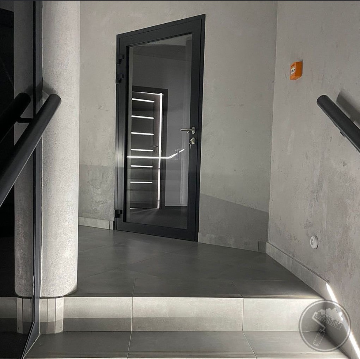 Szare płytki podłogowe w korytarzu biurowym z kolumną i szklanymi drzwiami z ciemną ramą, przez które widać oświetlone drzwi w głębi.