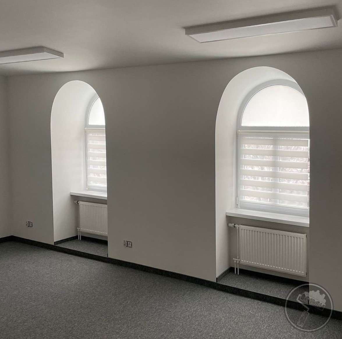 Wnętrze biura z dwoma oknami w kształcie łuku, świeżo pomalowane ściany na biało, szary dywan i białe grzejniki.