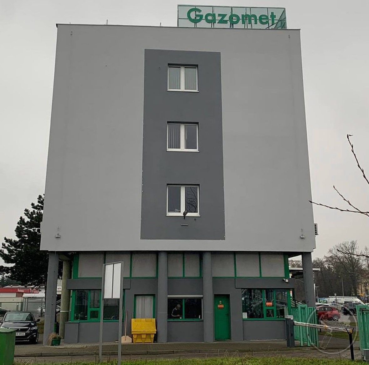 Szary budynek biurowy z zielonymi akcentami i napisem 'Gazomet' na dachu, widziany z poziomu ulicy w pochmurny dzień.