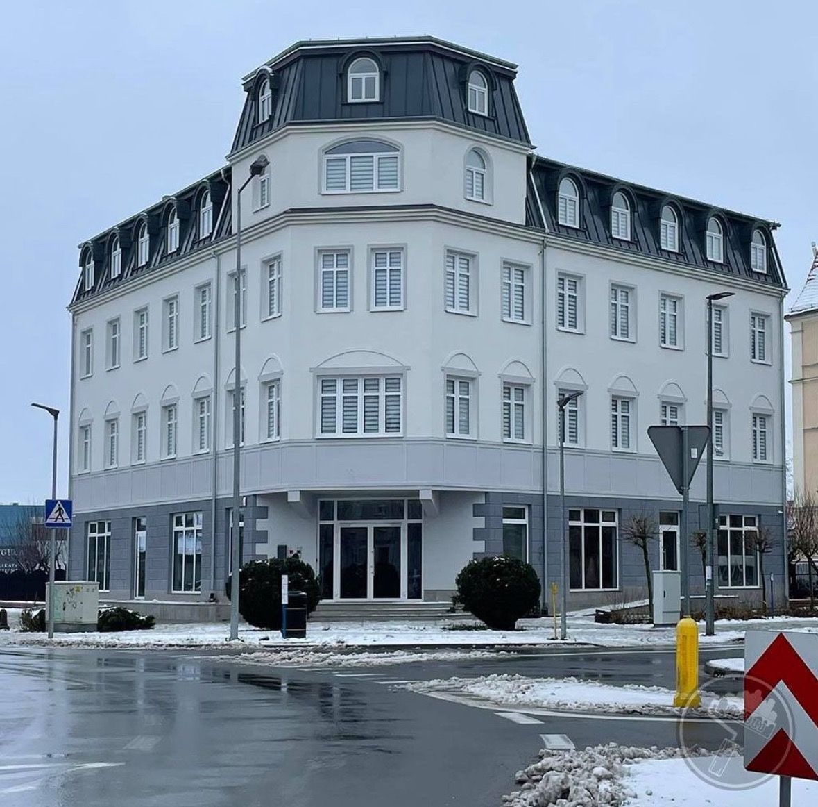 Odnowiony, trzypiętrowy budynek biurowy z szarym dachem mansardowym i białymi ścianami, widoczny z poziomu ulicy w pochmurny dzień, z elementami krajobrazu miejskiego i resztkami śniegu.