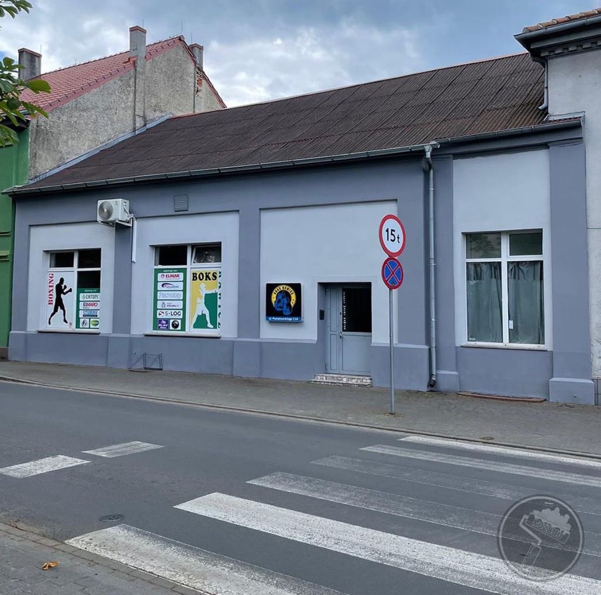 Odnowiony budynek z szarą elewacją, widoczne okna z reklamami, znak drogowy ograniczenia tonażu i przejście dla pieszych na pierwszym planie.