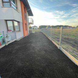 MM - Przygotowany teren pod trawnik, świeżo wyrównana ziemia obok nowoczesnego domu z balkonem i metalowym ogrodzeniem panelowym.