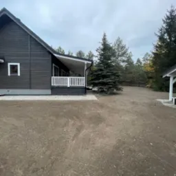 Przygotowany teren pod trawnik z rolki, widoczny dom z ciemnego drewna i posadzona choinka.