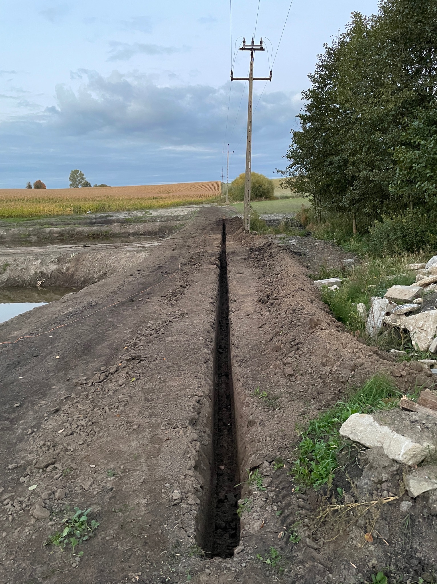 Wąski, głęboki rów wykopany w ziemi na tle pola uprawnego i słupów energetycznych, widoczne ślady opon i kamienie na brzegu wykopu.