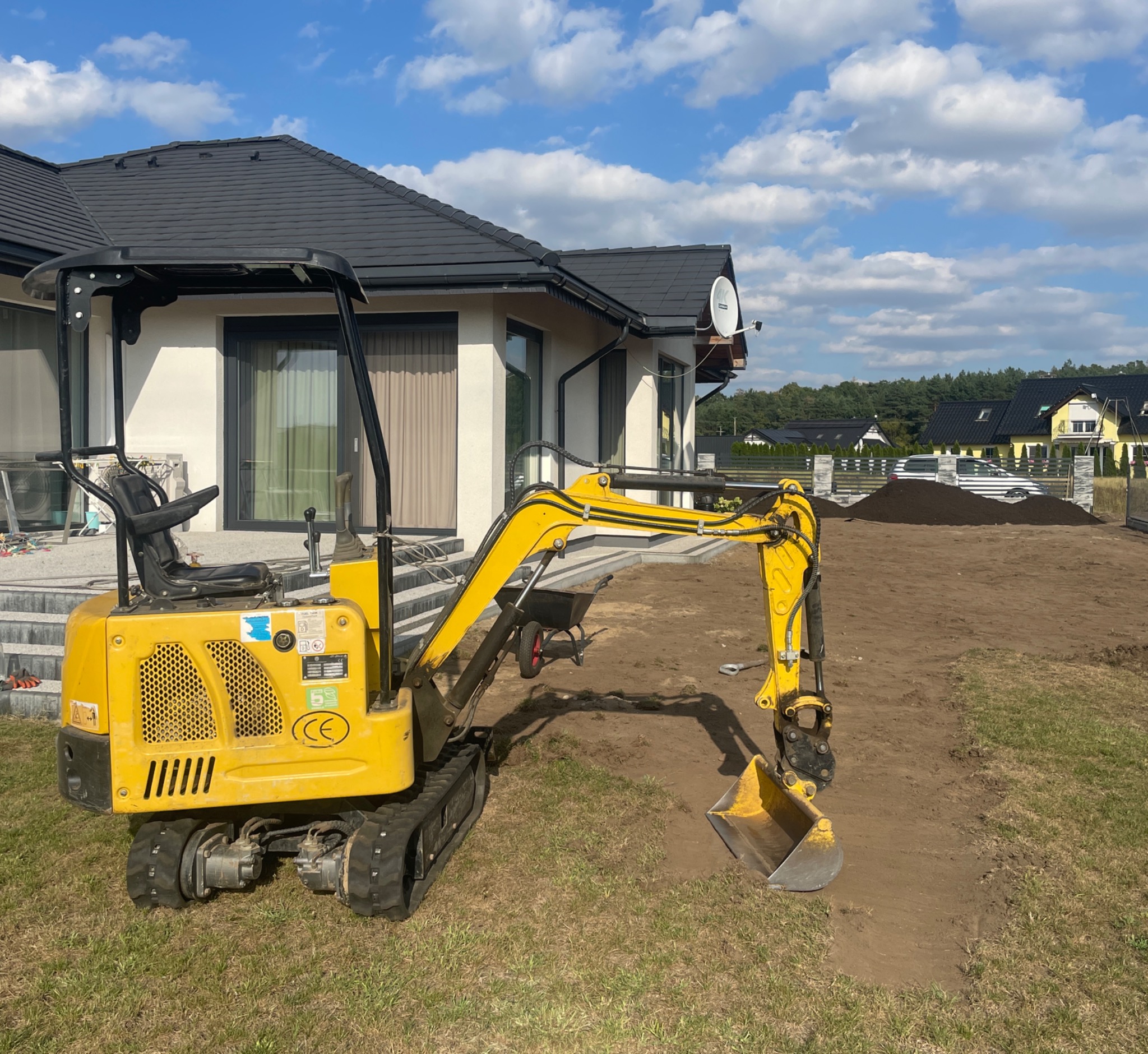 Żółta mini koparka na gąsienicach, z wykopaną bruzdą w trawie, obok nowoczesnego domu z ciemnym dachem i stertą ziemi w tle.