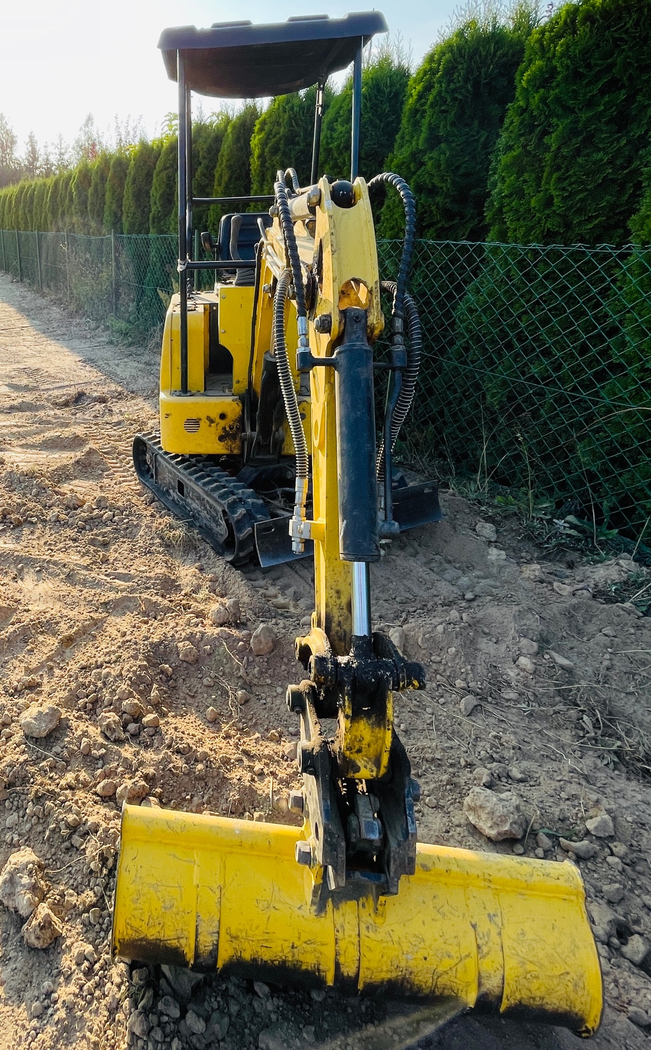 Żółta minikoparka z łyżką na ziemi, widoczna na tle zielonego żywopłotu i siatki ogrodzeniowej, w trakcie prac ziemnych.