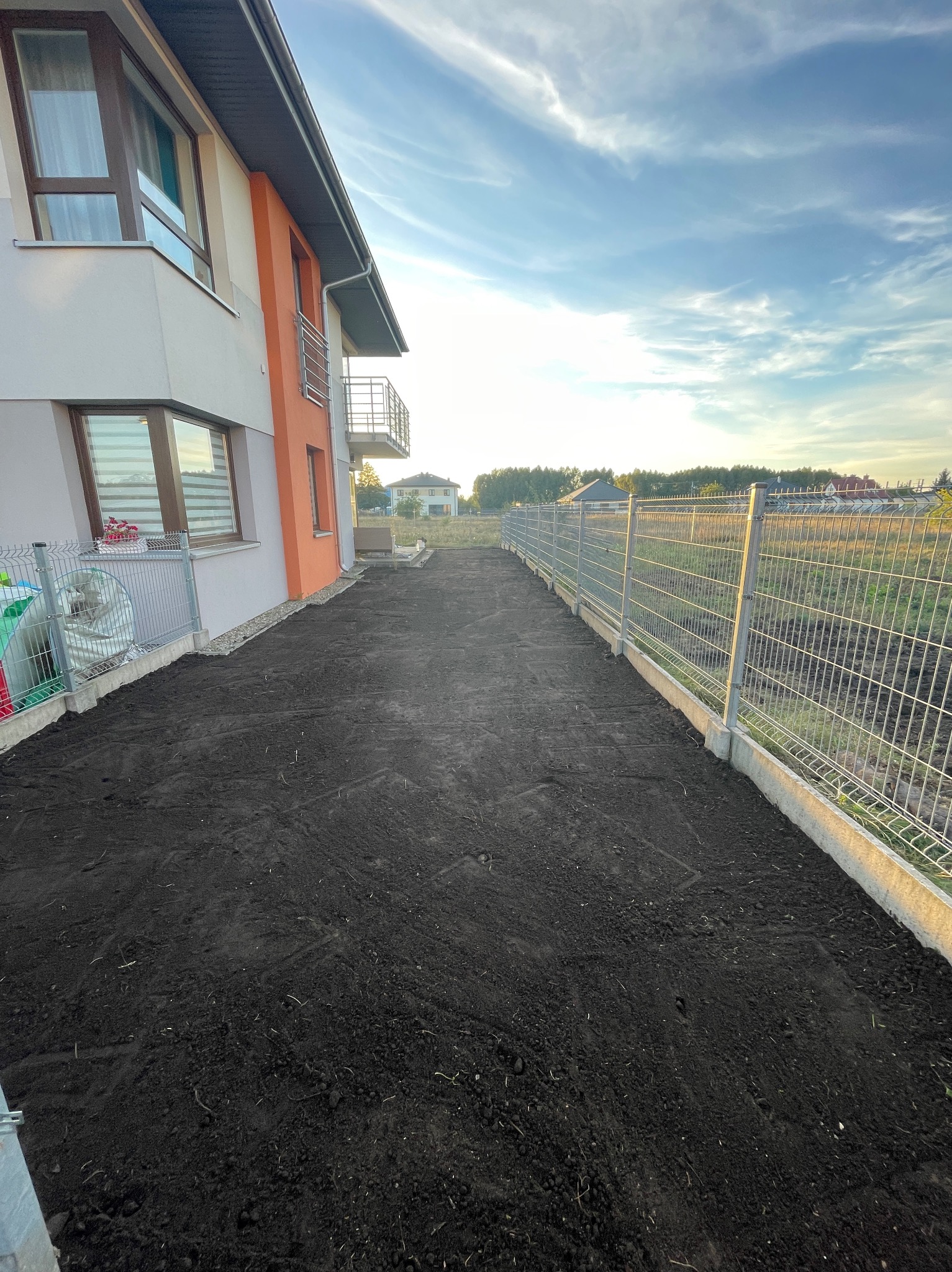 Przygotowany teren pod trawnik, świeżo wyrównana ziemia obok nowoczesnego domu z balkonem i metalowym ogrodzeniem panelowym.