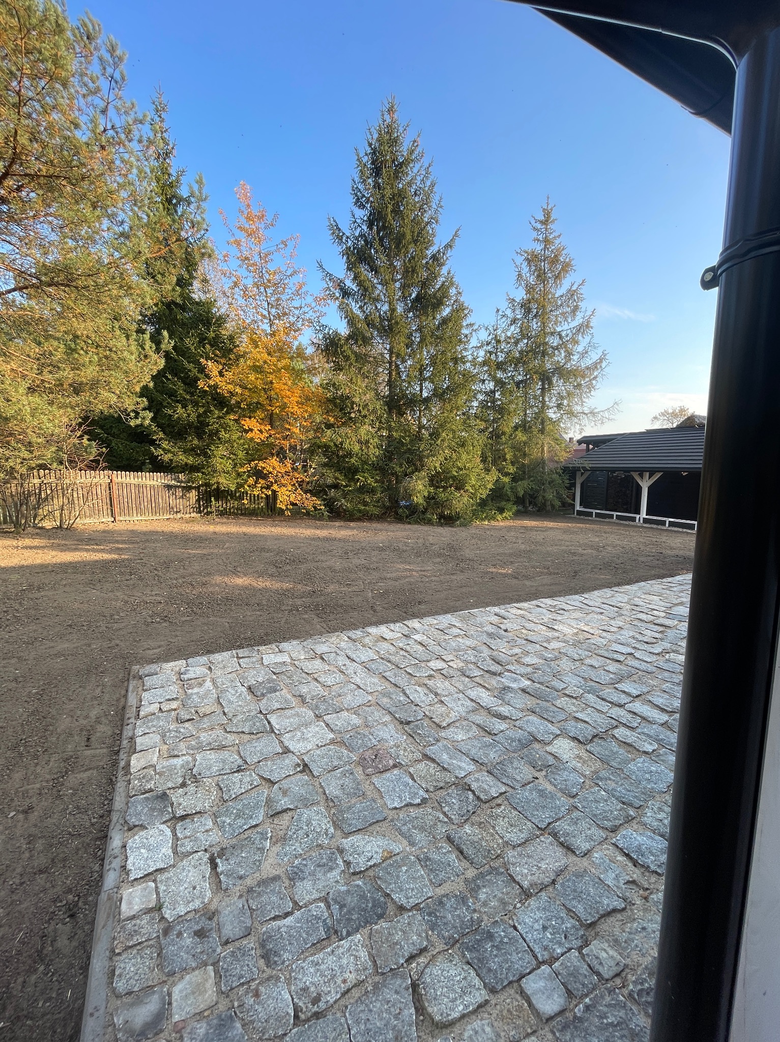 Przygotowany teren pod trawnik z widoczną kostką granitową, ogrodzeniem i zabudową ogrodową w tle, słoneczny dzień.