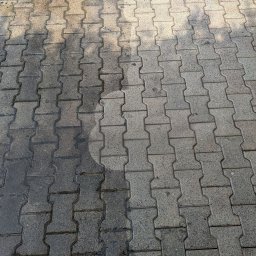 Marami Rafał Olszewski - Fragment chodnika z kostki brukowej, gdzie widoczna jest różnica w czystości między umytą a nieumytą częścią, z okrągłym śladem po czyszczeniu pod ciśnieniem.