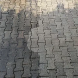 Fragment chodnika z kostki brukowej, gdzie widoczna jest różnica w czystości między umytą a nieumytą częścią, z okrągłym śladem po czyszczeniu pod ciśnieniem.