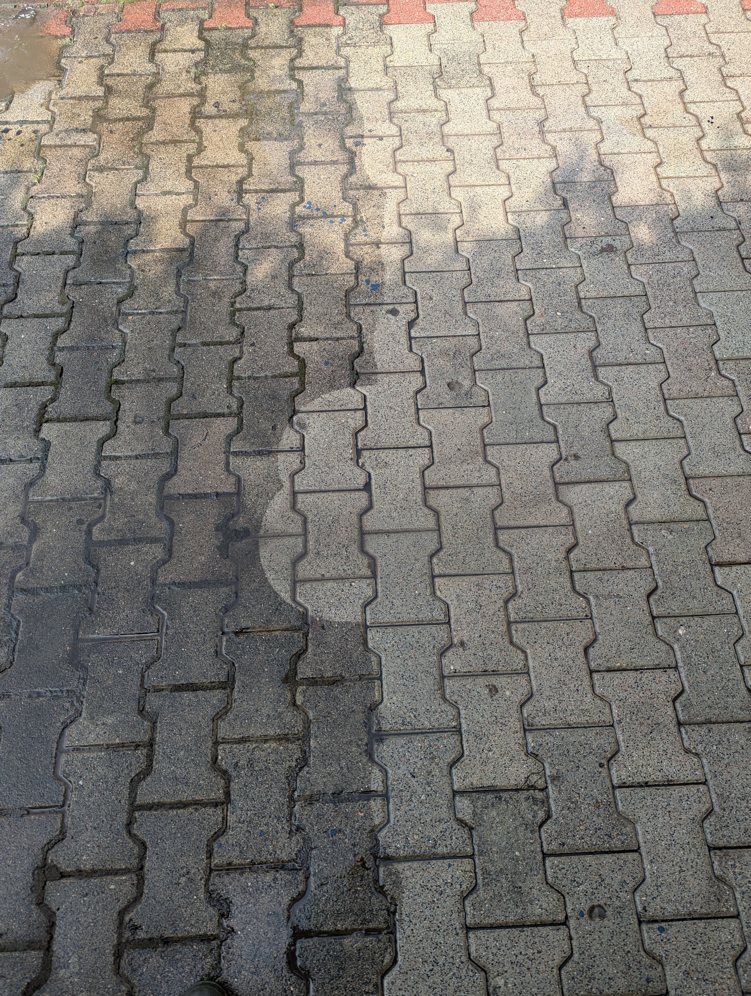 Fragment chodnika z kostki brukowej, gdzie widoczna jest różnica w czystości między umytą a nieumytą częścią, z okrągłym śladem po czyszczeniu pod ciśnieniem.