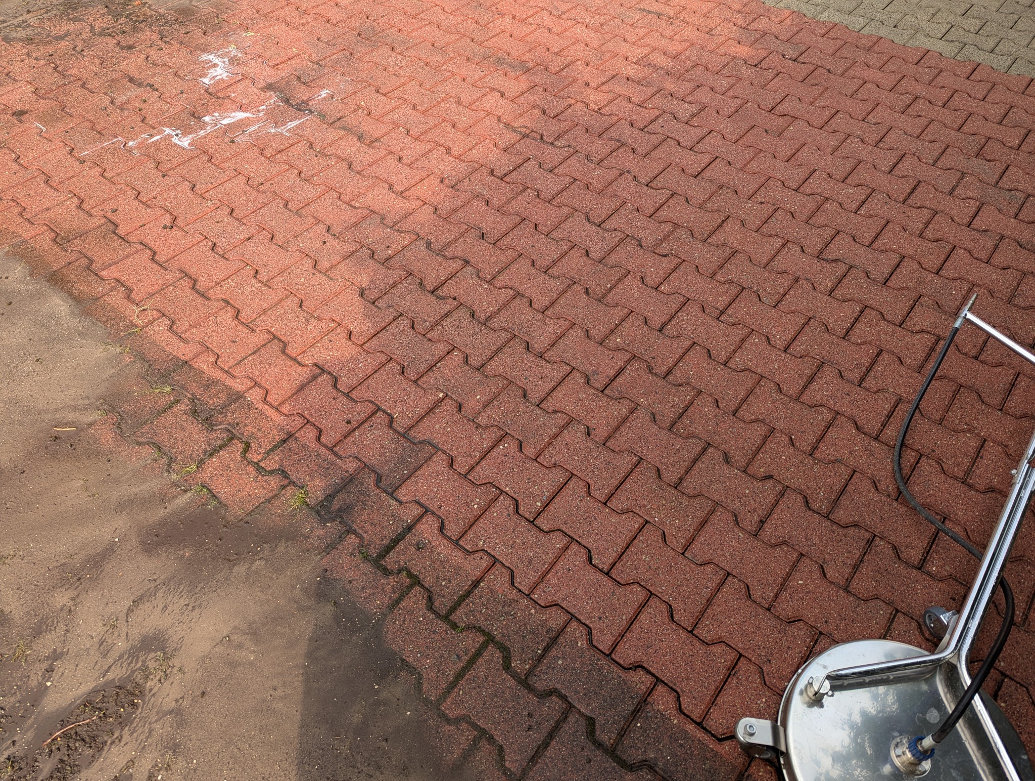 Częściowo umyta czerwona kostka brukowa z widocznym urządzeniem do czyszczenia pod ciśnieniem, ukazująca różnicę między czystą a zabrudzoną powierzchnią.