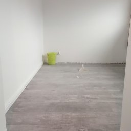 MAPKIM remonty - Wnętrze po remoncie z nową podłogą z płytek imitujących drewno, widoczny zielony kosz na śmieci i rurka miedziana pod sufitem.