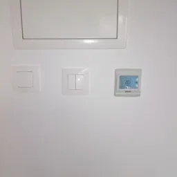Biała ściana z klapą rewizyjną, pojedynczym i podwójnym włącznikiem światła oraz termostatem pokazującym temperaturę 20.5 stopni Celsjusza.
