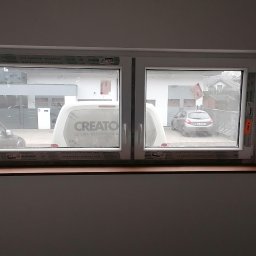 MAPKIM remonty - Widok z wnętrza na dwa okna PCV z brązowym parapetem, na zewnątrz zaparkowane samochody i van z napisem CREATO.