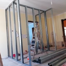 MAPKIM remonty - Wnętrze w trakcie remontu z metalową konstrukcją ścian działowych, drewnianą drabiną z plamami farby, profile podłogowe, mężczyzna w czapce obserwujący postęp prac, widoczne fragmenty starej...