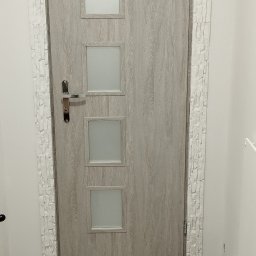 MAPKIM remonty - Szare drzwi wewnętrzne z czterema kwadratowymi szybkami, obramowane dekoracyjną białą okładziną imitującą kamień, na tle paneli podłogowych w odcieniach szarości.